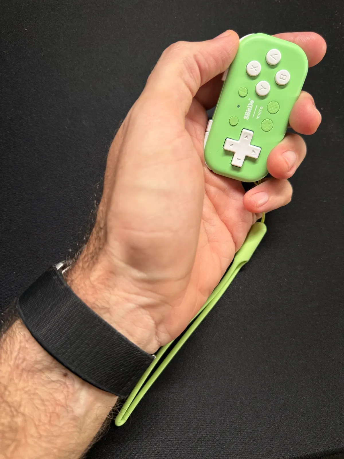 The 8BitDo Micro, a small green game controller, held vertically in the left hand, with a wrist strap.